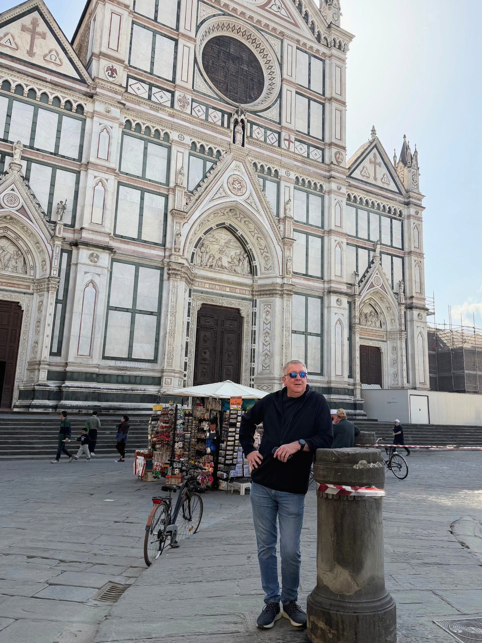 A man wearing sunglasses and a black jacket stands next to a street vendor stand in front of the Florence Cathedral in Florence, Italy. The cathedral's ornate facade with marble details is visible in the background. Several people are walking on the square, and a bicycle is parked nearby.