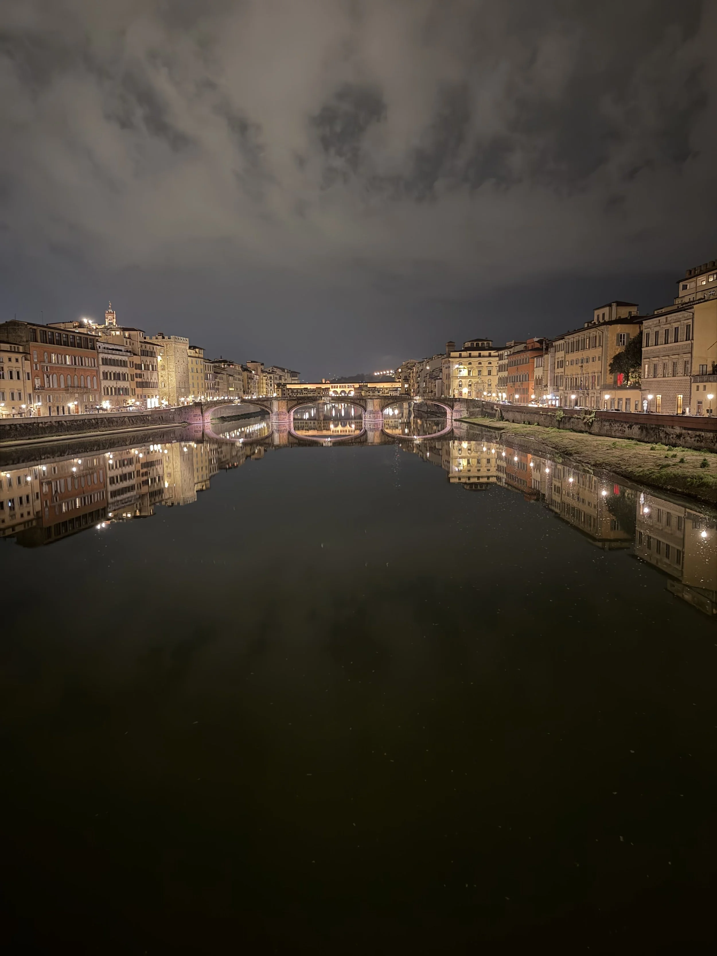 Nighttime view of a city canal with reflections of buildings and a bridge illuminated with purple lights.