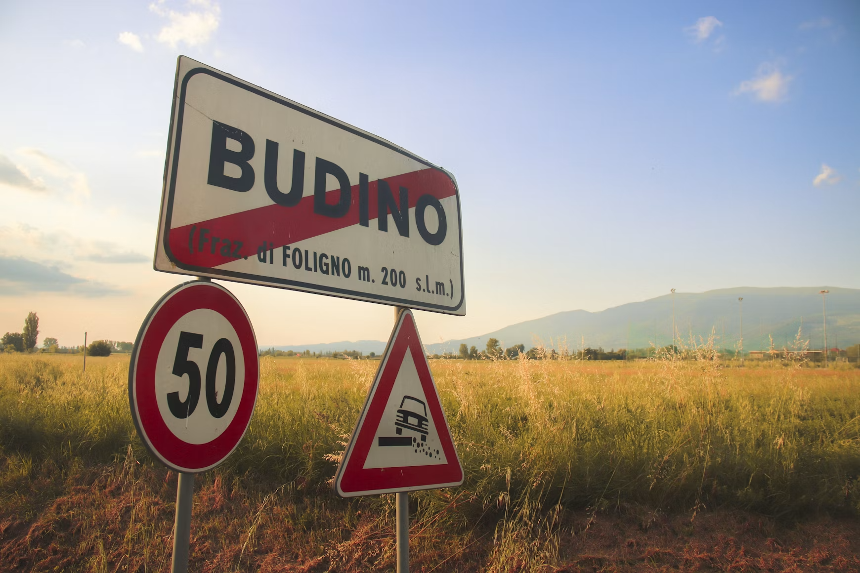 Road signs indicating a speed limit of 50 km/h and a warning for loose gravel on the road, with a town name sign for Budino, Italy, in a rural landscape at sunset.