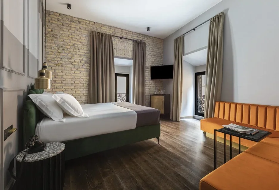Modern hotel room featuring a bed with green headboard, brick accent wall, window with beige curtains, orange sofa, small black side table with magazines, and wall-mounted TV.