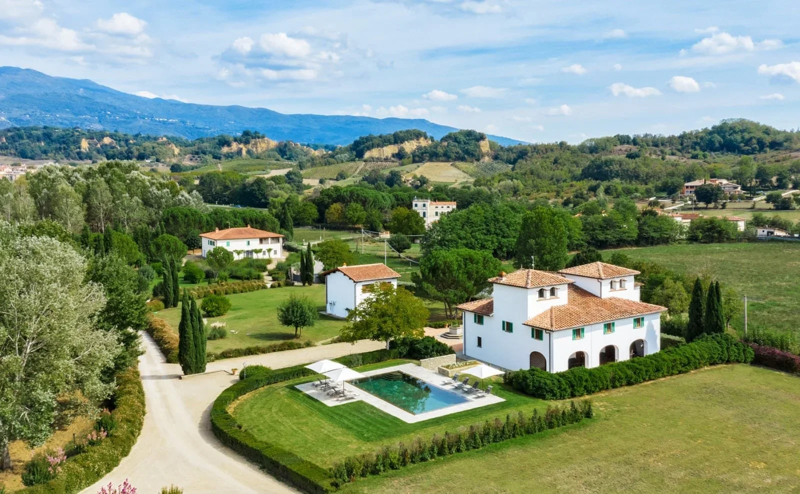 Scenic view of a Mediterranean-style estate with white villas, a swimming pool with lounge chairs, surrounded by lush greenery and rolling hills in the background