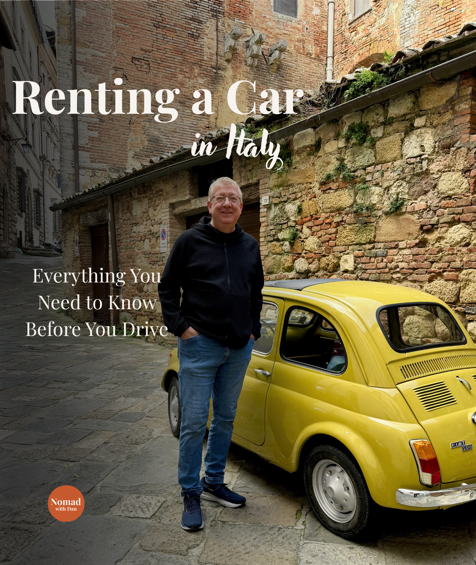 A man in a black jacket and blue jeans standing next to a bright yellow vintage Fiat 500 car on a cobblestone street in Italy with brick buildings in the background.