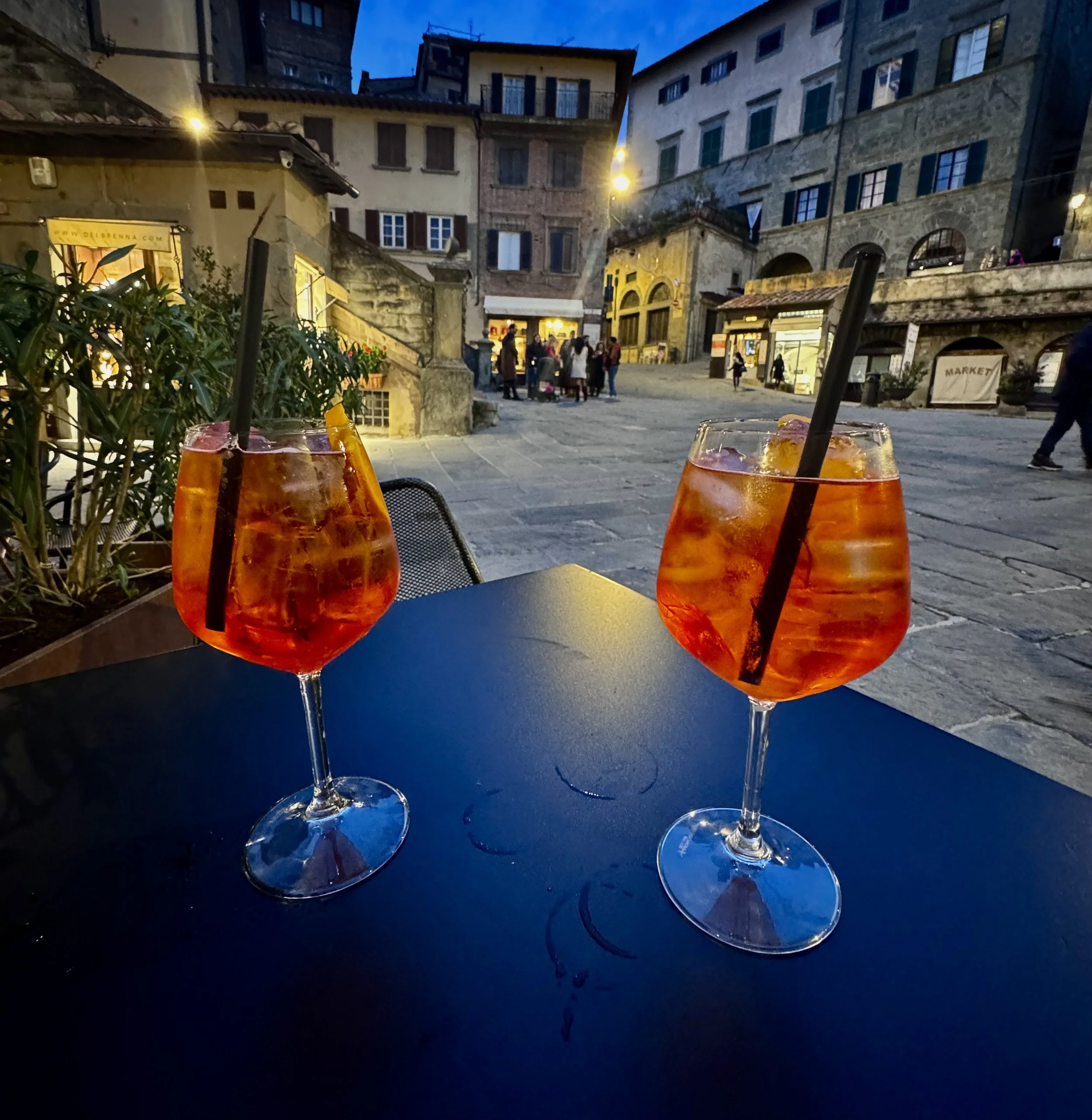 Two glasses of orange-colored drink with ice and straws on a dark table, with a city square and people walking in the background during the evening.