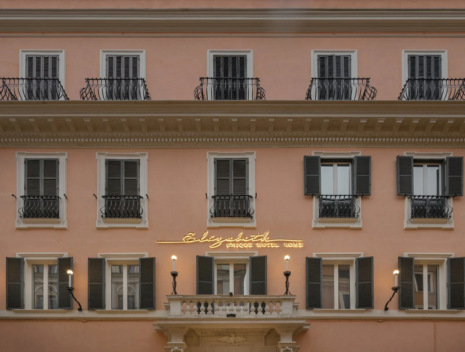 The facade of a pink hotel with several windows with black shutters and balconies; the hotel name "Elizabeth" is illuminated in yellow lights, with a small balcony below it and vintage street lamps lighting the entrance area.