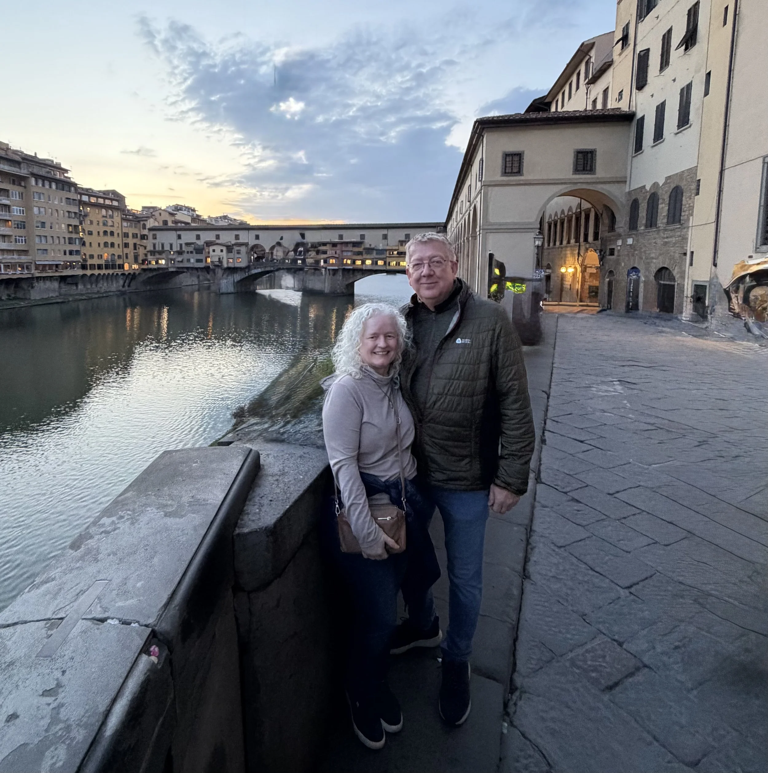 A smiling couple standing on a stone sidewalk next to a river during sunset in Florence, Italy, with historic buildings and a bridge in the background.