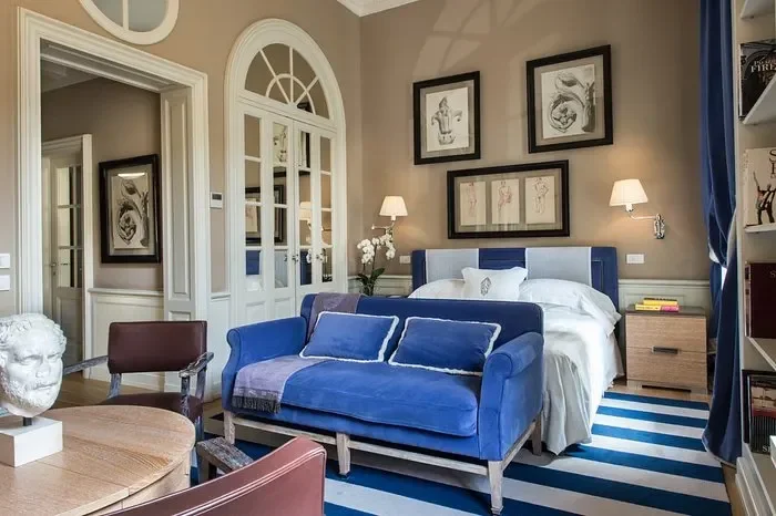 A bedroom with beige walls, a bed with white bedding, blue cushions, and a blue headboard. There are two wall-mounted lamps, framed artwork above the bed, a bedside table with books, and a blue and white striped rug. A blue velvet loveseat is in front of the bed, and there's a wooden table with a bust sculpture in the foreground.