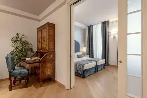 A bedroom seen through an open sliding door, featuring two beds with blue bedding, a window with blue curtains, and a lamp.