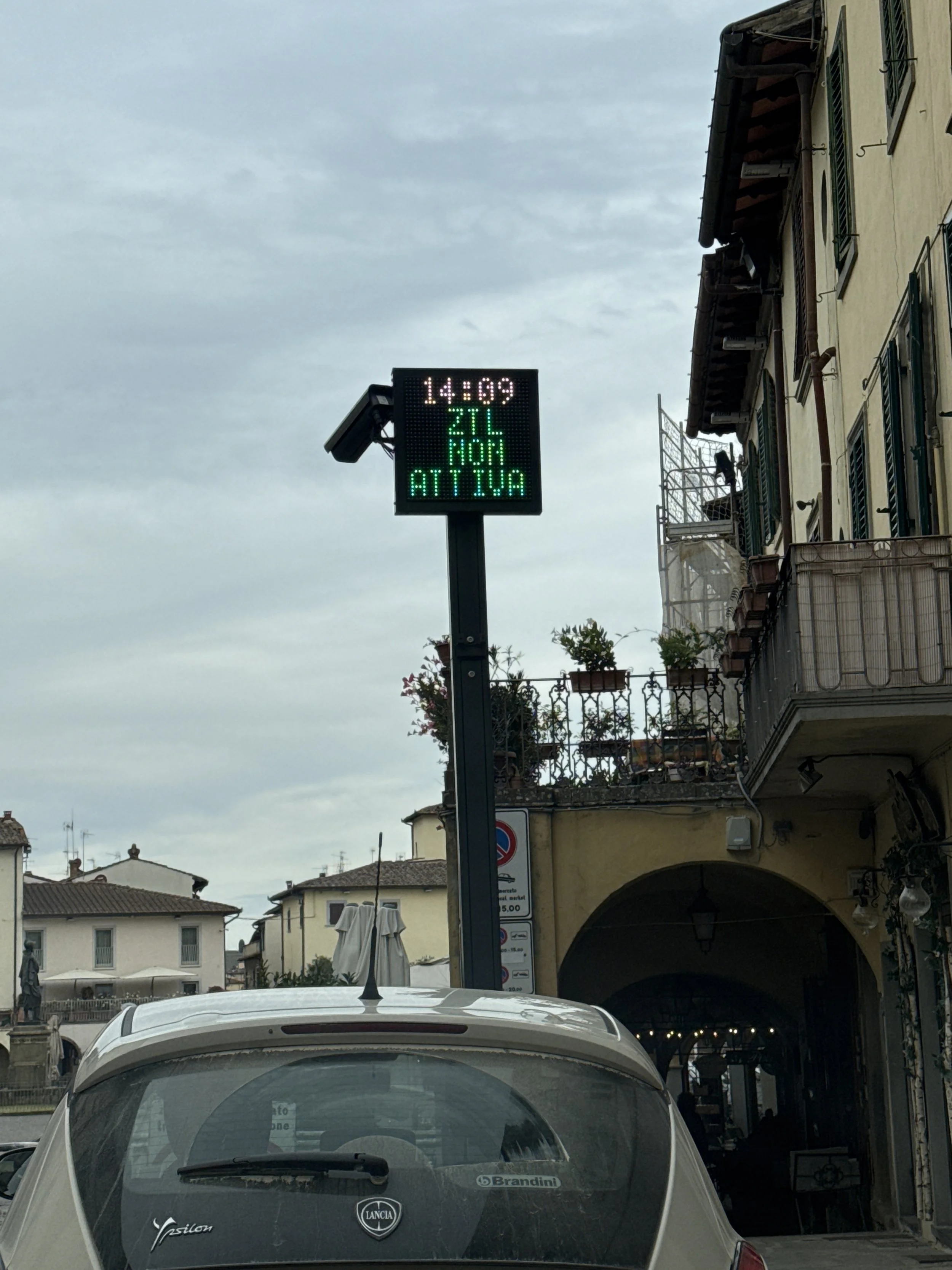View of a parking area with a digital sign displaying the time 14:09, temperature 27°C, and a message in Italian. Buildings with balconies and plants, some under construction, are visible in the background.