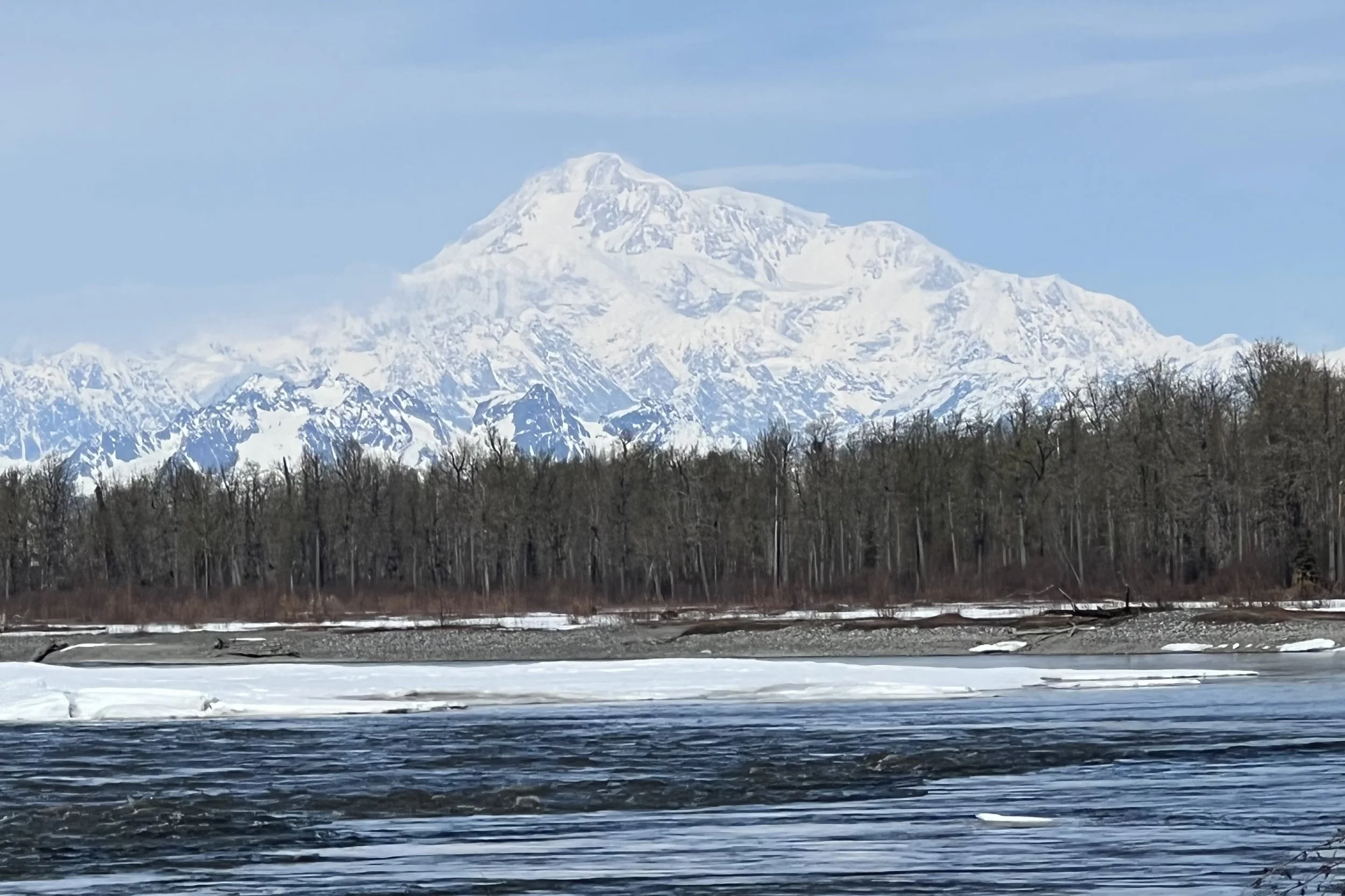 Snow-covered mountain range with a prominent peak in the background, a forest of leafless trees in the middle ground, and a partially frozen river in the foreground.