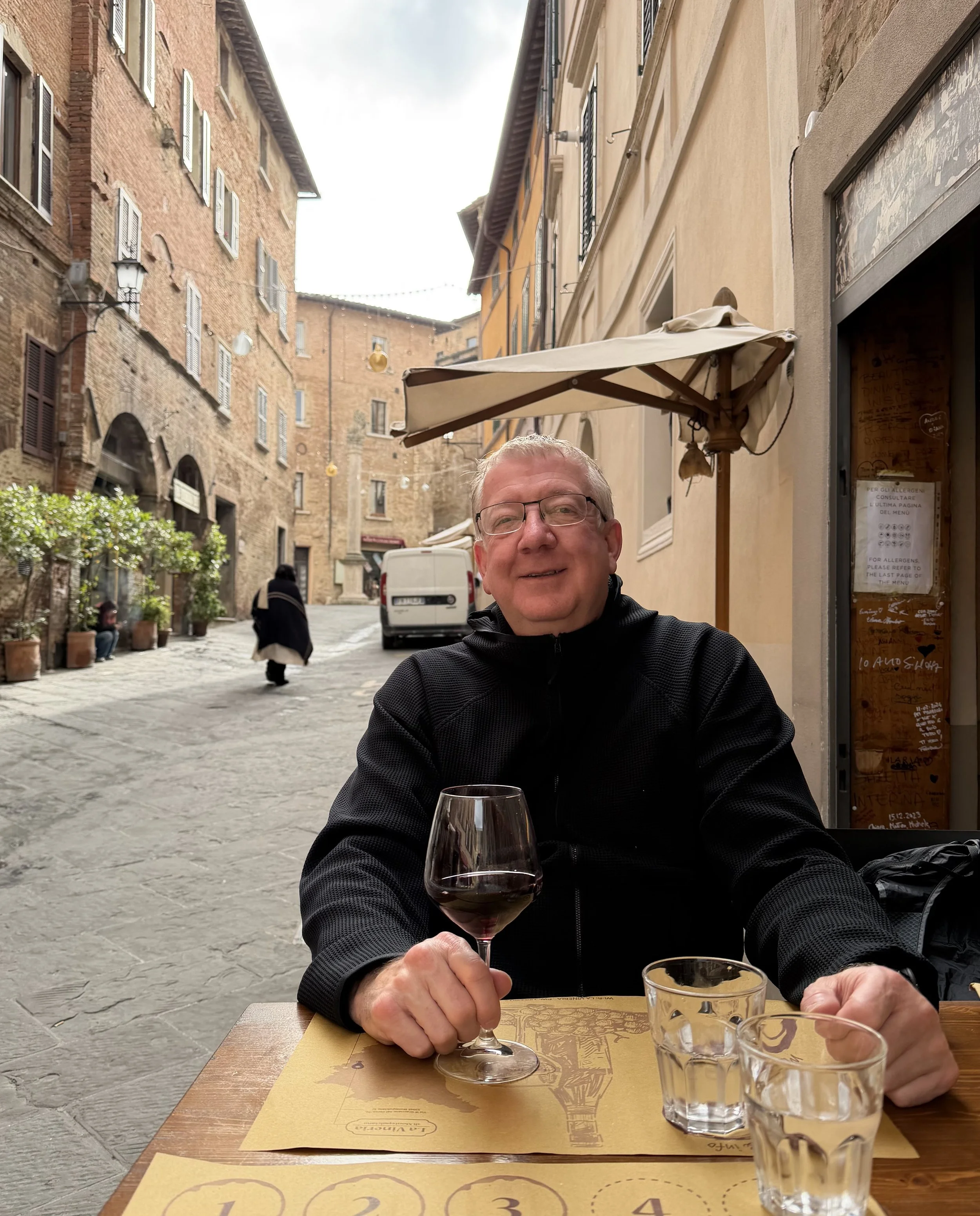 A man with glasses and a black jacket sitting at an outdoor restaurant table, holding a glass of red wine. Other glasses and a menu are on the table. The background shows a European street with brick buildings, potted plants, and pedestrians walking.