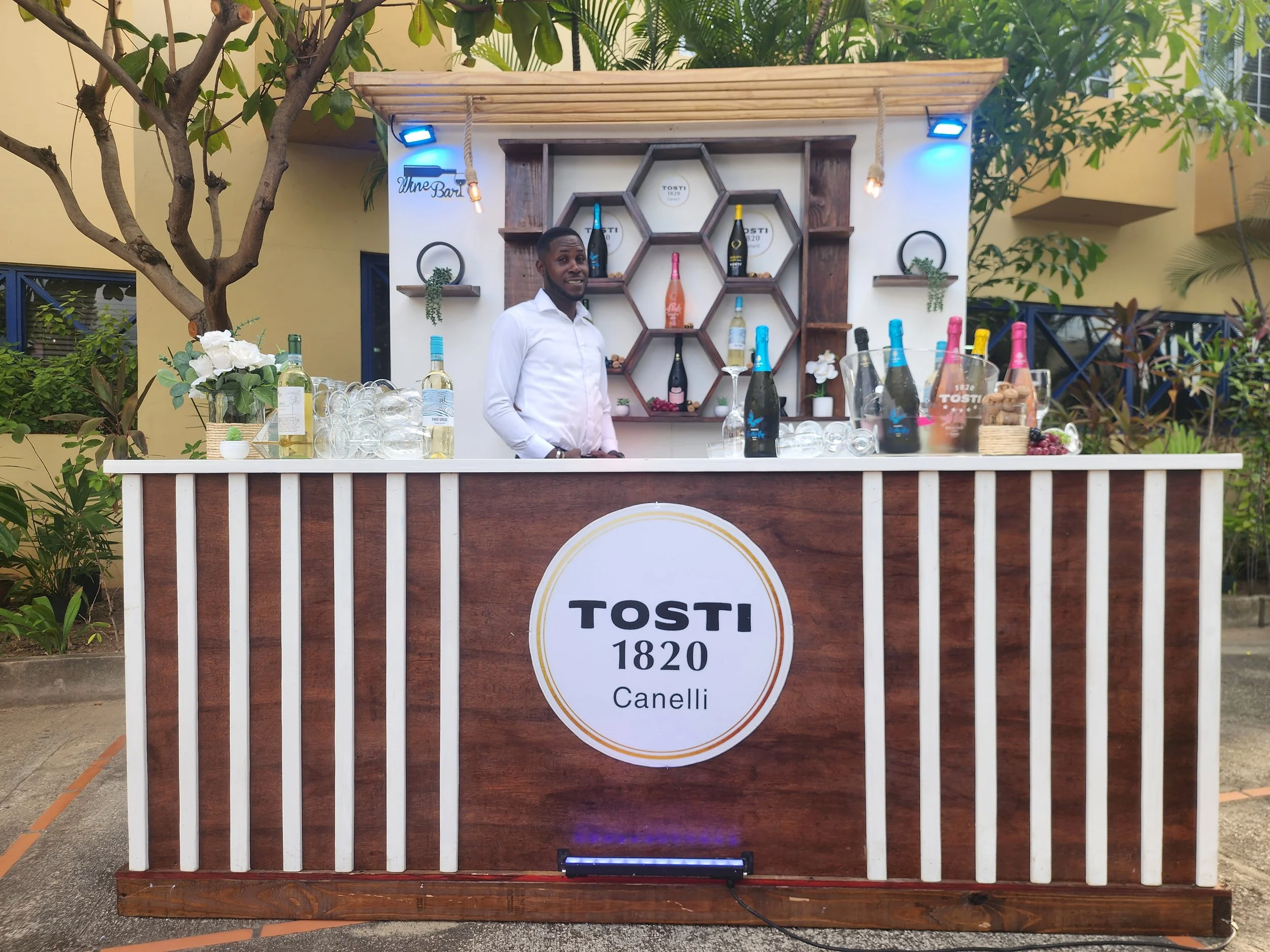 A man standing behind a bartender's counter at a Tosti 1820 Canelli wine booth outdoors, with bottles of wine, ice, and glasses on the counter, surrounded by greenery and yellow building walls.