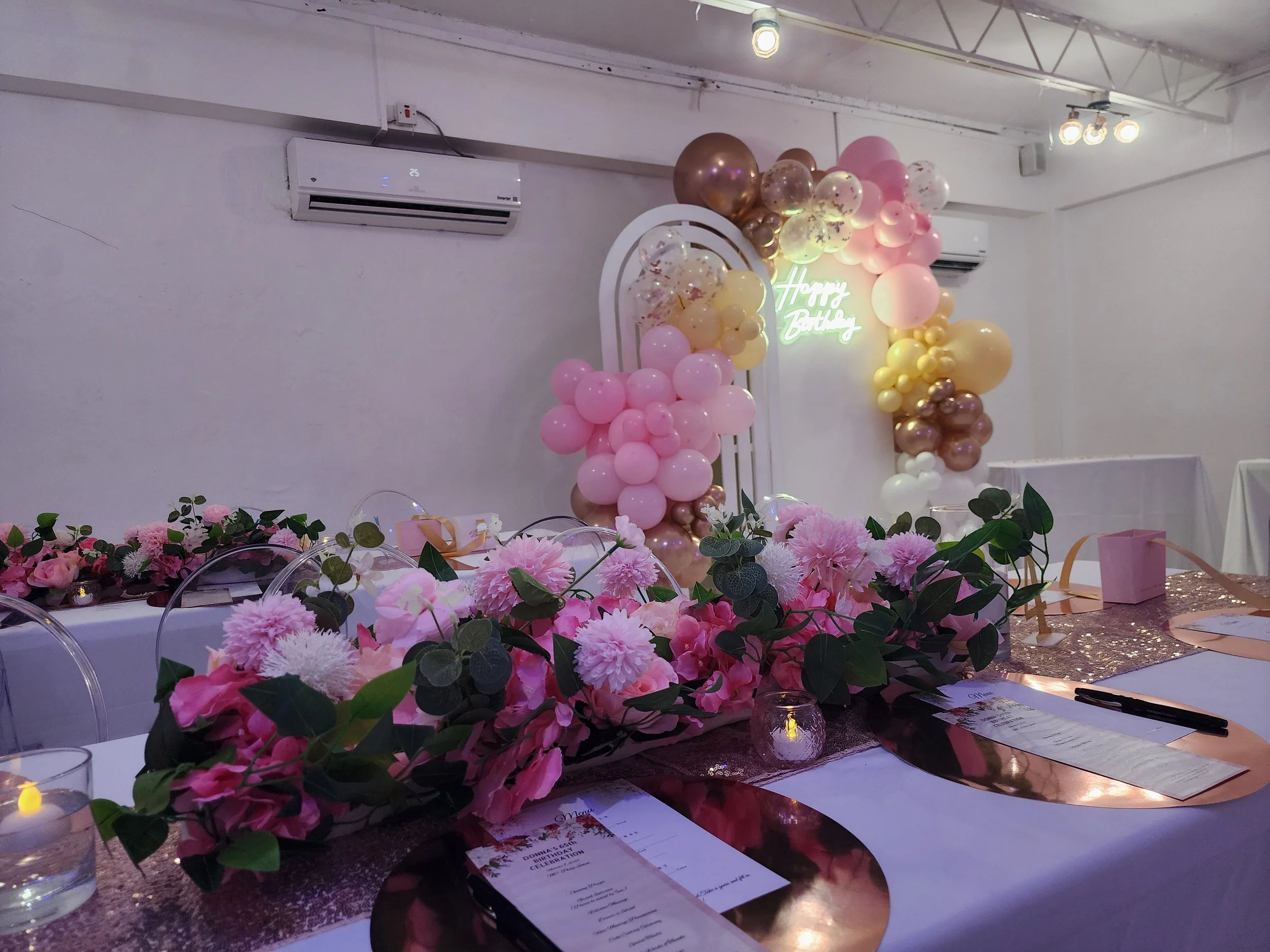Decorated banquet table with pink and white floral arrangements, candles, and place cards; pink, gold, and white balloon arch and a neon sign reading "Happy Birthday" in the background