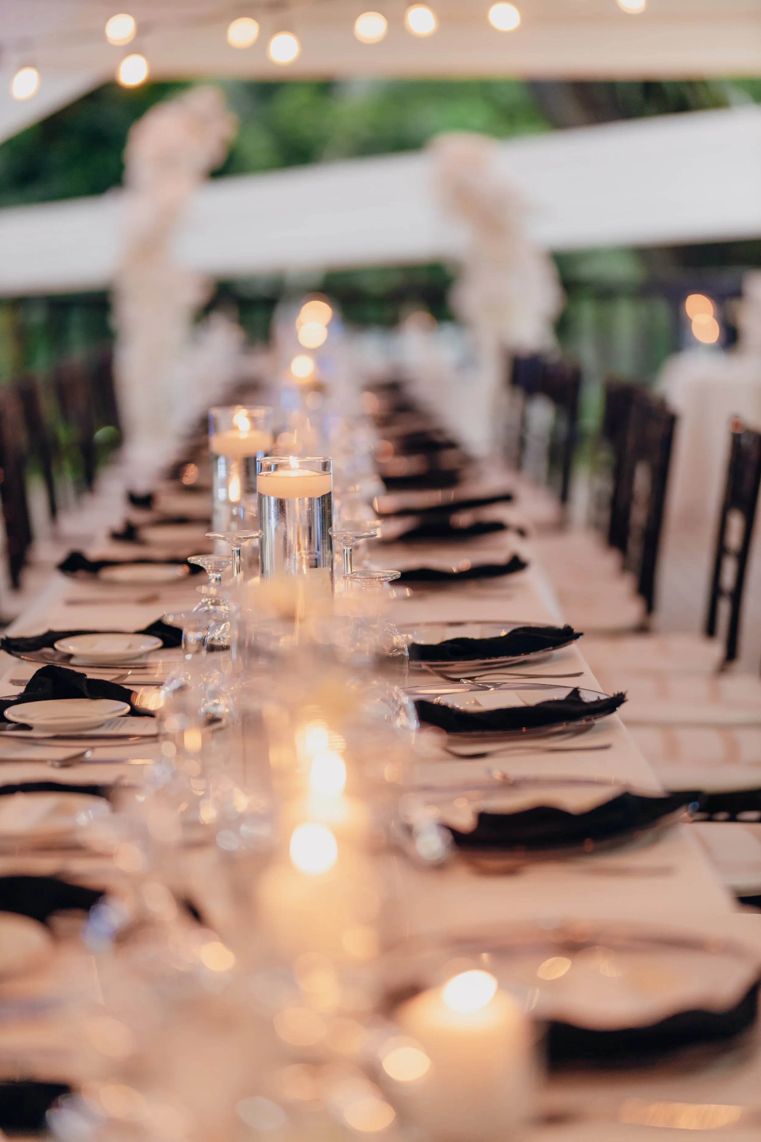 Chic black & white wedding tablescape. Dinner table set with black napkins, glassware, plates, and lit candles at an elegant outdoor event.