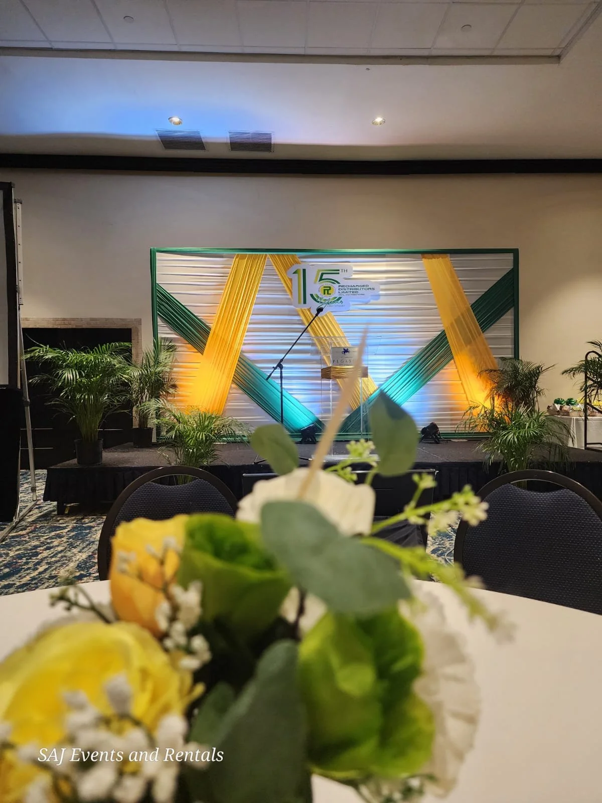Event stage decorated with yellow and teal ribbons, with a 15th anniversary logo, plants, and a flower arrangement in the foreground.