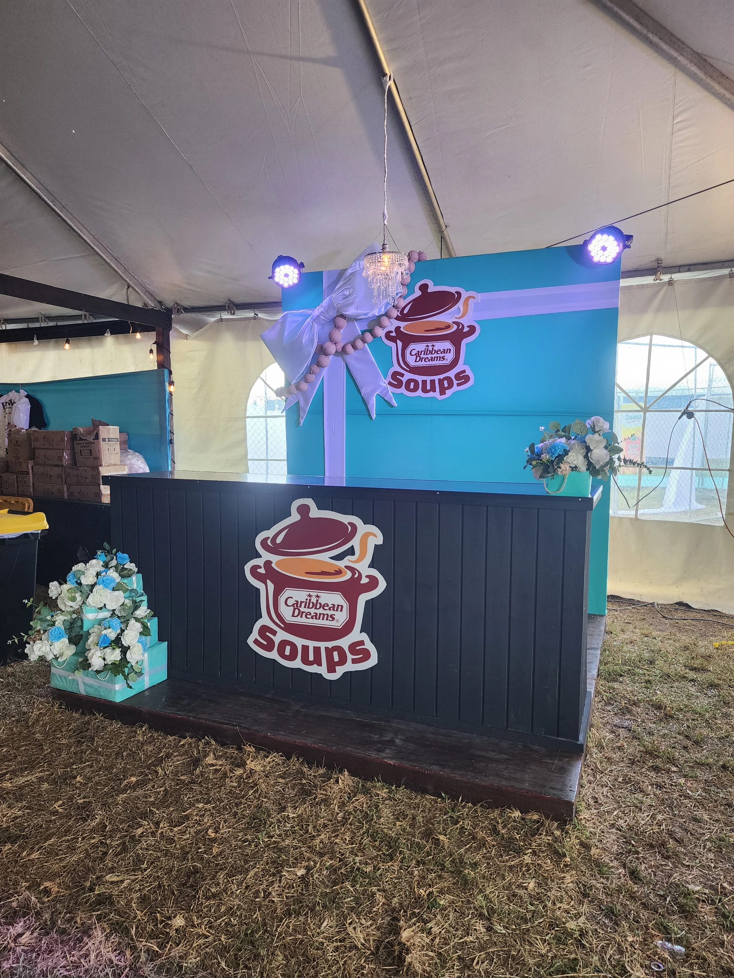 Decorated food booth with Caribbean Dreams soups sign, blue background, floral arrangements, and hanging chandelier inside a tent.