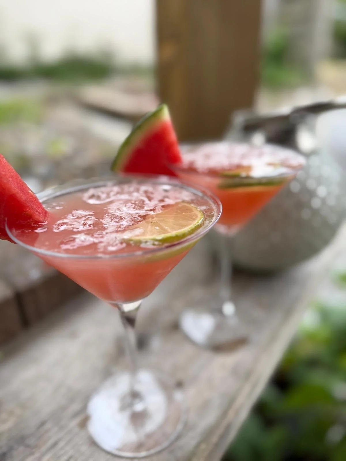 Two pink cocktails with lemon and watermelon garnishes on a wooden surface outdoors.