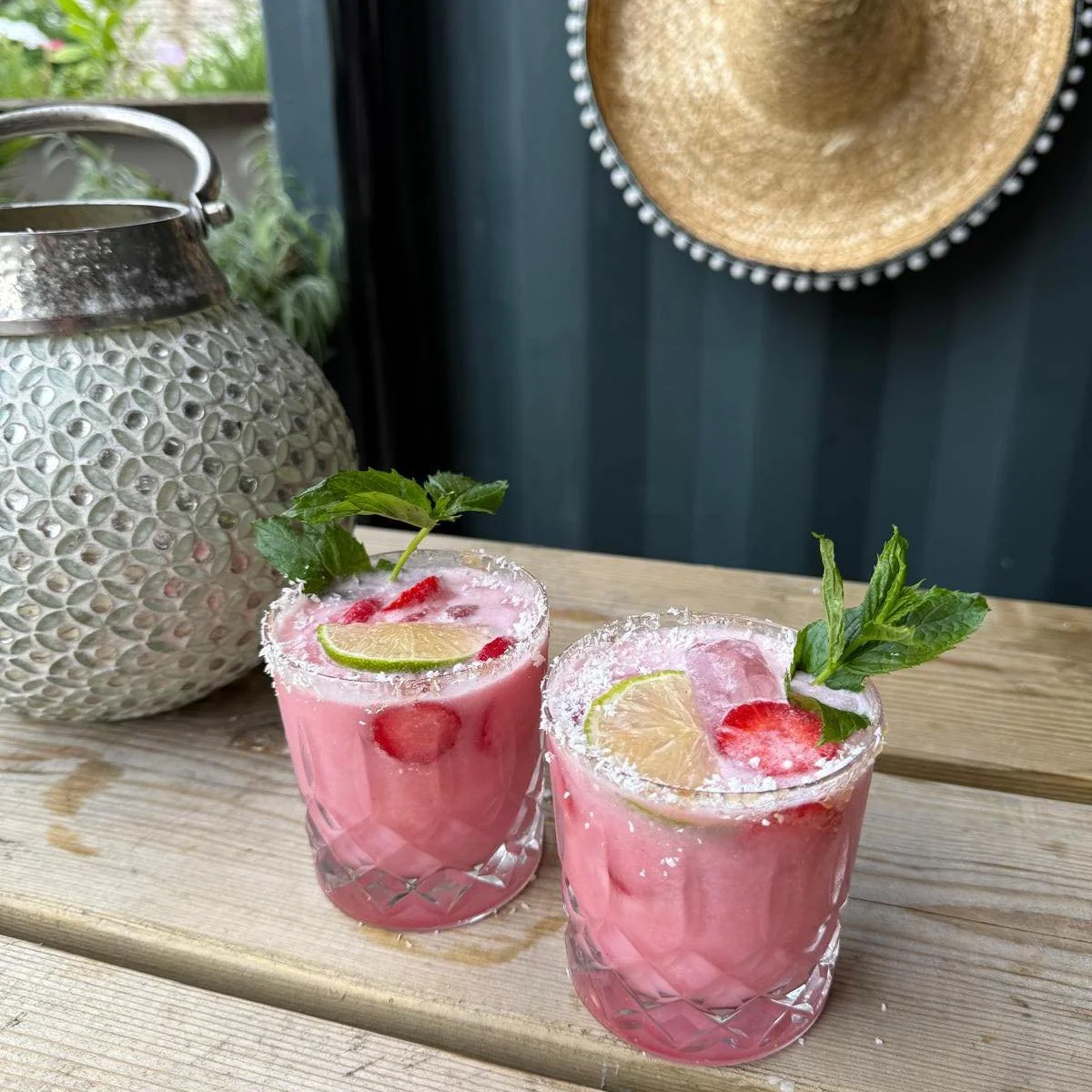 Two pink cocktails garnished with lime slices, strawberry pieces, mint leaves, and shredded coconut, placed on a wooden surface near a decorative lantern and a hat hanging on a dark green wall.
