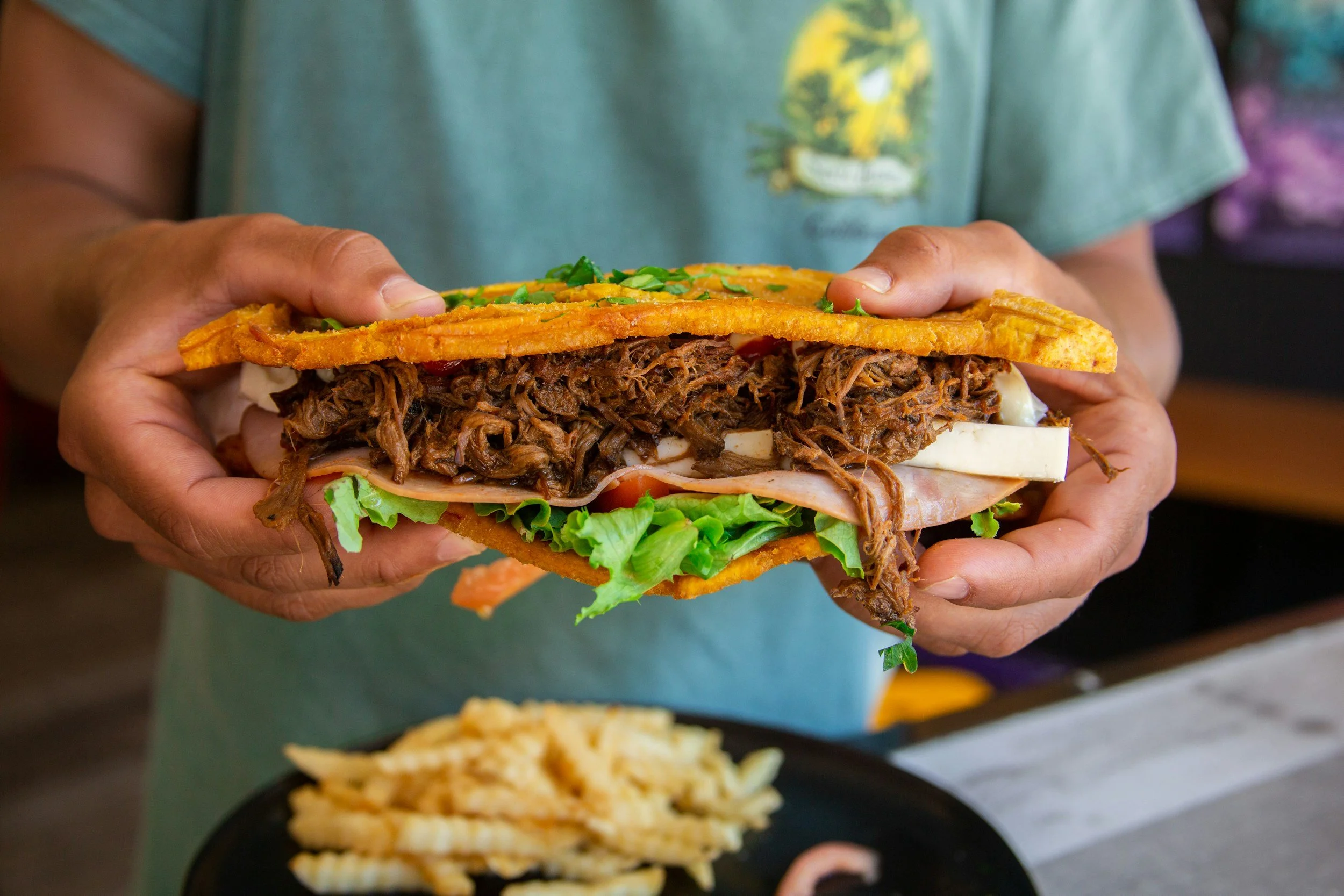 Person holding a large street food item with shredded beef, lettuce, tomato, cheese, and two crispy waffle cones