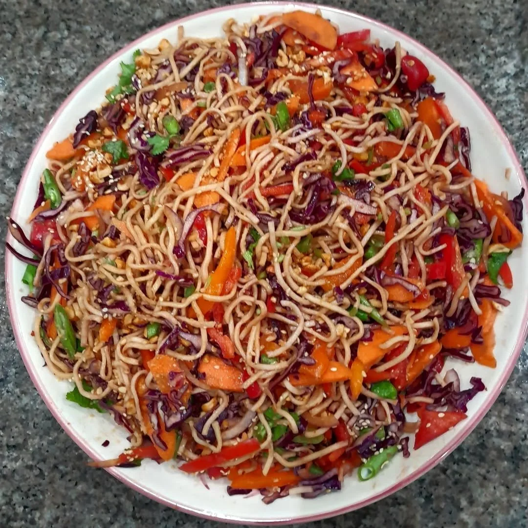 A plate of Asian-style noodle salad with shredded carrots, red bell peppers, purple cabbage, chopped green onions, and sesame seeds on top.