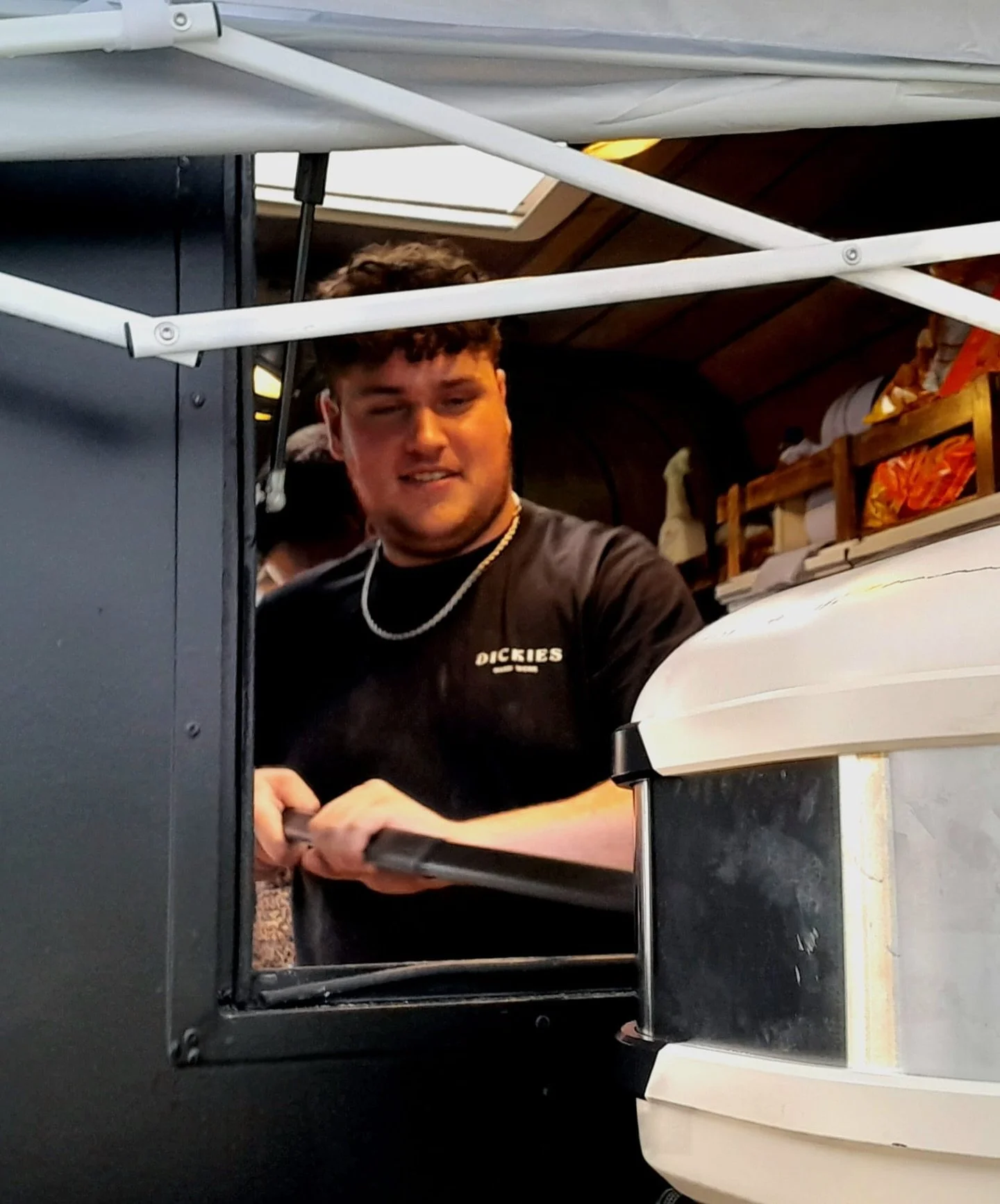 A young man with short dark hair wearing a black t-shirt with 'DICKIES' written on it, standing in a kitchen or food service area, cooking wood fired pizza, leaning on a counter handle and looking at the camera.