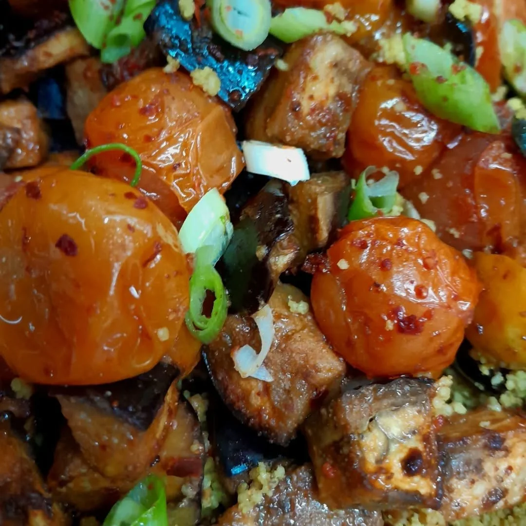 Close-up of a dish with cherry tomatoes, diced eggplant, chopped green onions, and tofu cubes in a sauce with red chili flakes.