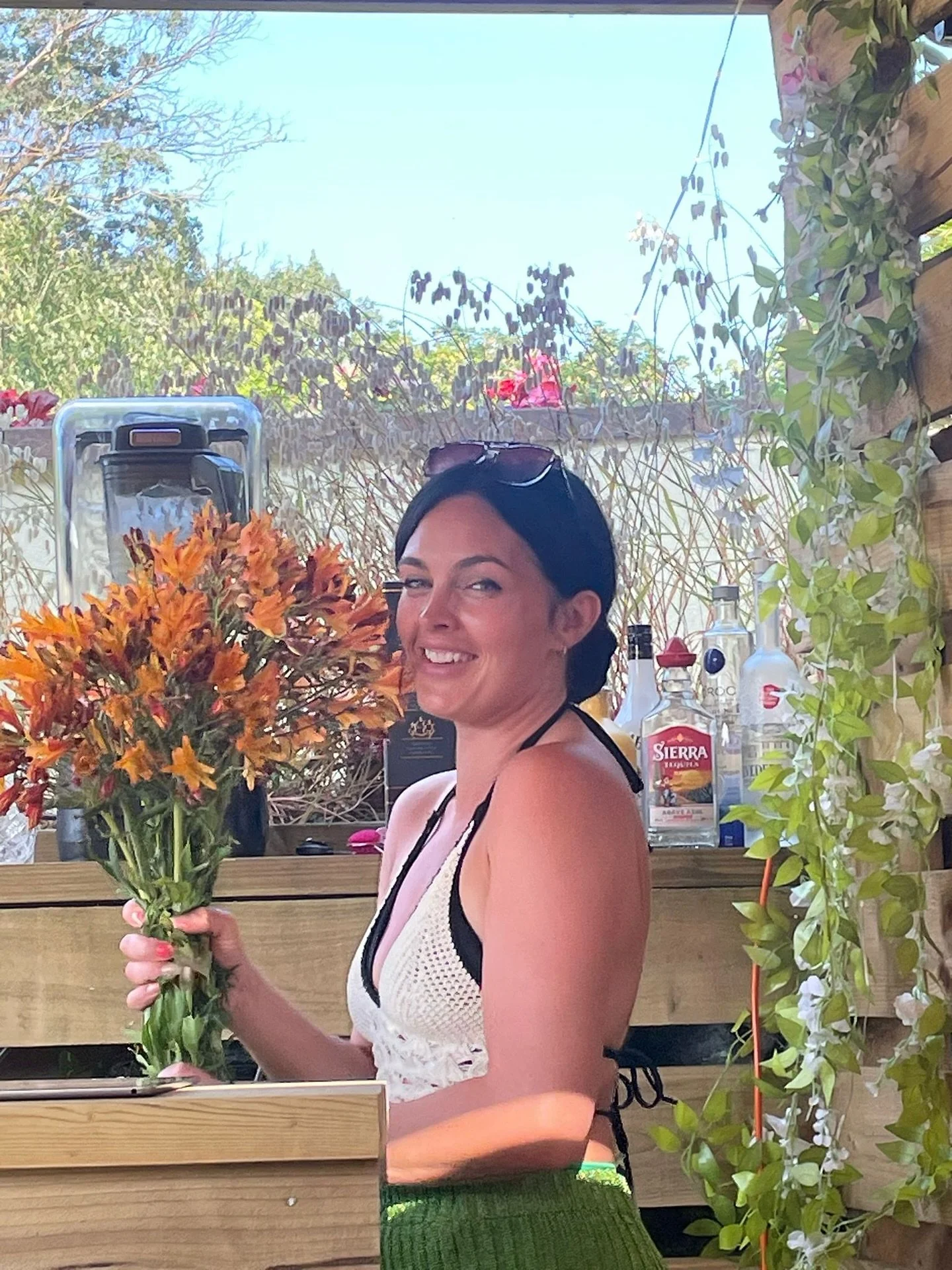 A smiling woman holding a bouquet of orange flowers in an outdoor setting with plants and bottles in the background.