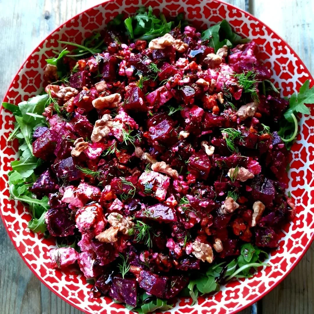 A salad with roasted beets, walnuts, fresh herbs, and mixed greens in a patterned red and white bowl on a wooden surface.