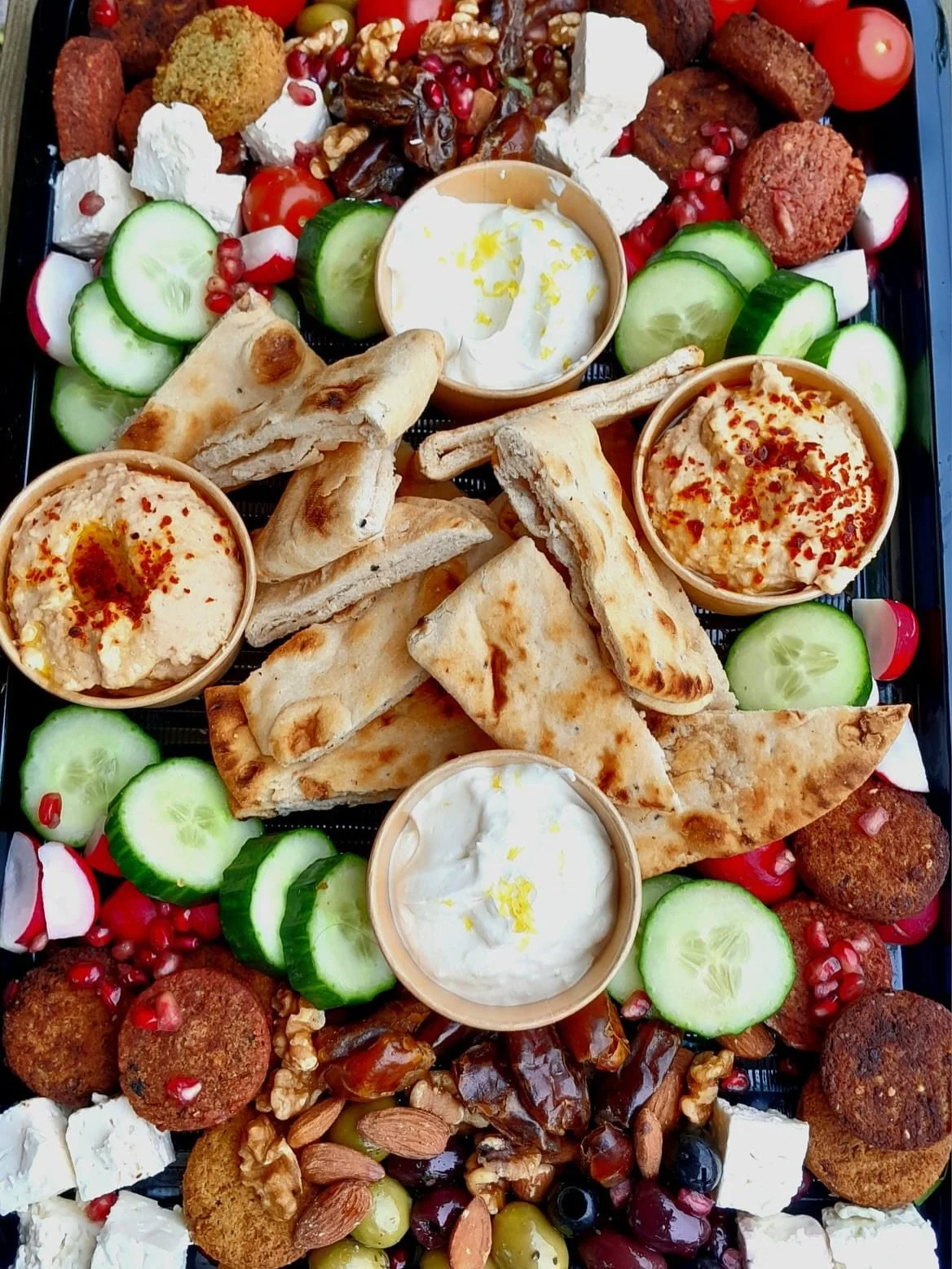 Assorted Middle Eastern appetizers on a platter including hummus with red pepper flakes, yogurt with lemon zest, pita bread triangles, cucumber slices, cherry tomatoes, falafel, stuffed grape leaves, cheese cubes, mixed nuts, pomegranate seeds, and radish slices.