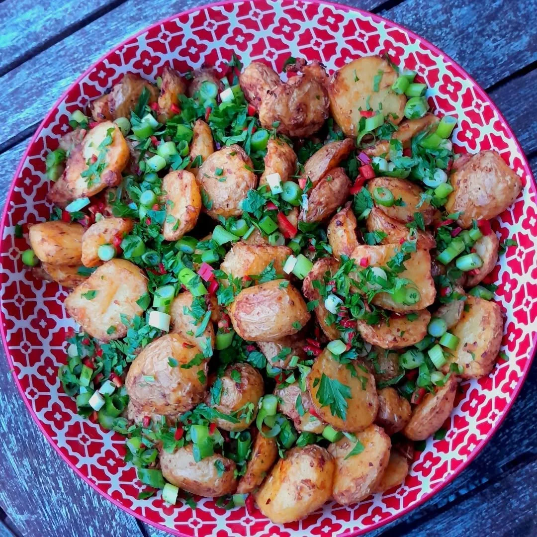 A red and white patterned bowl filled with roasted potato chunks garnished with chopped green onions and parsley, placed on a wooden surface.