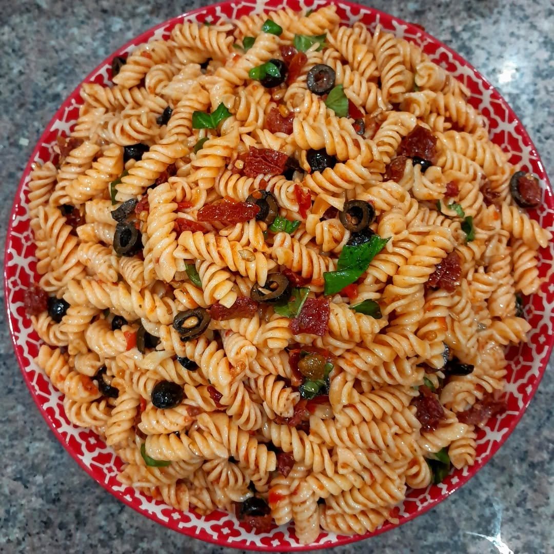 A red bowl filled with cooked fusilli pasta mixed with tomato, black olives, green onions, and sauce, placed on a speckled gray countertop.
