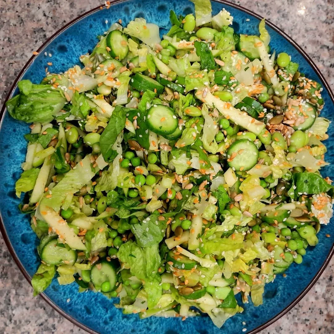 A colorful salad in a blue bowl containing chopped lettuce, cucumber slices, green peas, mixed seeds, and sesame seeds.