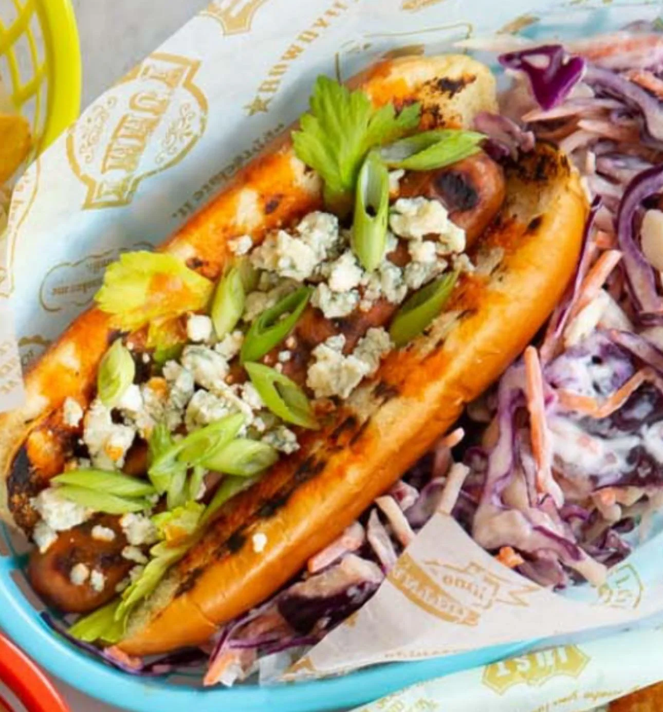 Hot dog with grilled sausage, topped with crumbled blue cheese and chopped green onions, served with side of coleslaw.