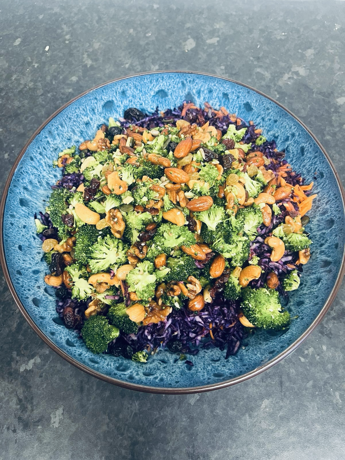 A colorful salad in a blue bowl containing broccoli, purple cabbage, shredded carrots, almonds, walnuts, and dried cranberries.