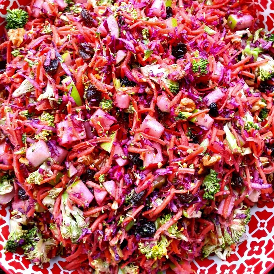 Colorful slaw salad with shredded carrots, purple cabbage, broccoli, and raisins on a decorative white and red plate.