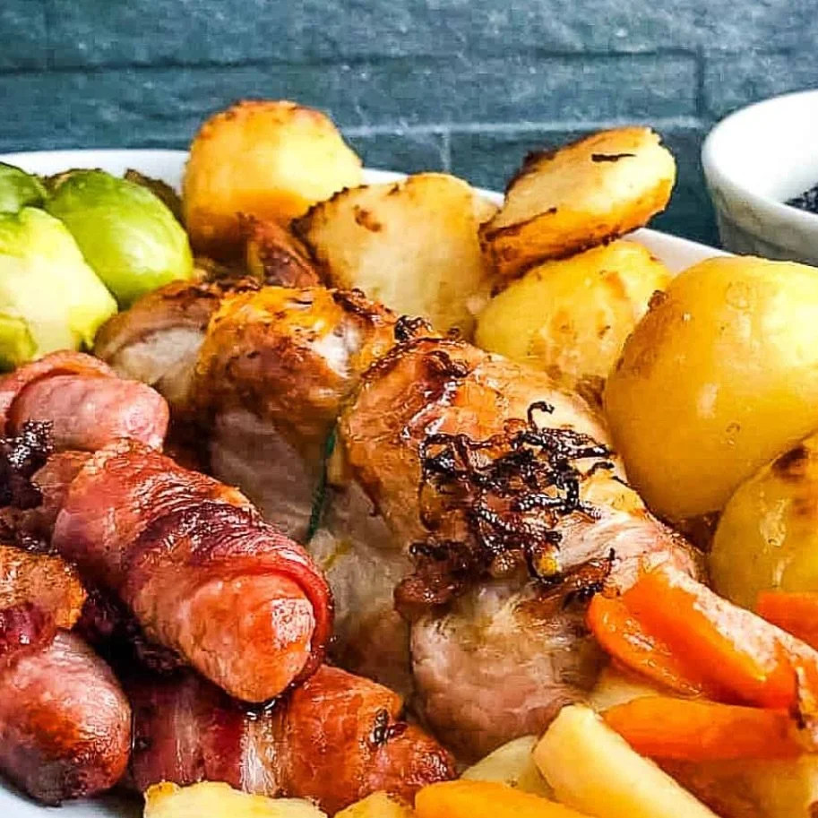 Close-up of roasted meat, sausages wrapped in bacon, roasted potatoes, and vegetables on a white plate.
