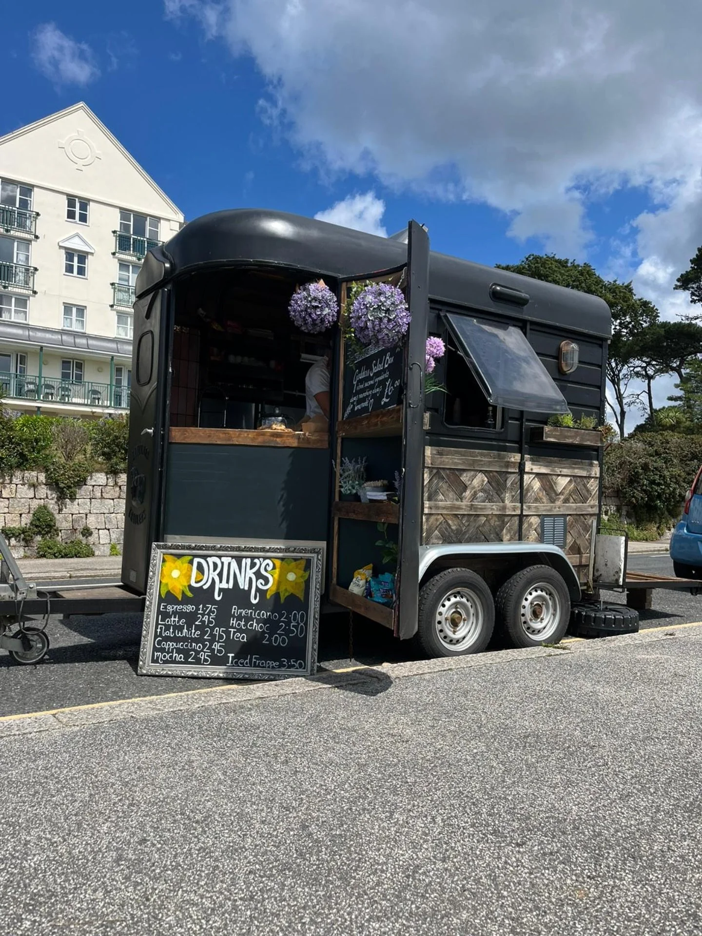 A black mobile coffee stand parked on the street with flowers hanging outside and a menu board listing various coffee drinks and prices.