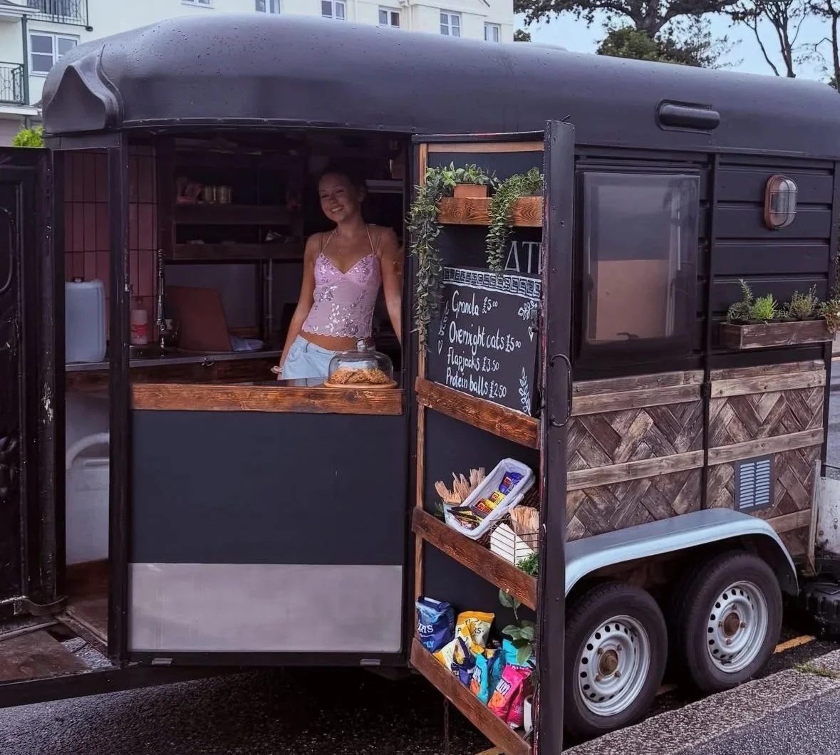 A woman selling snacks from a black food truck, with a chalkboard menu listing various food items like gnocchi, overnight oats, flapjacks, and protein balls. The truck has a rustic wood design, and there are snacks like chips displayed on a shelf outside.