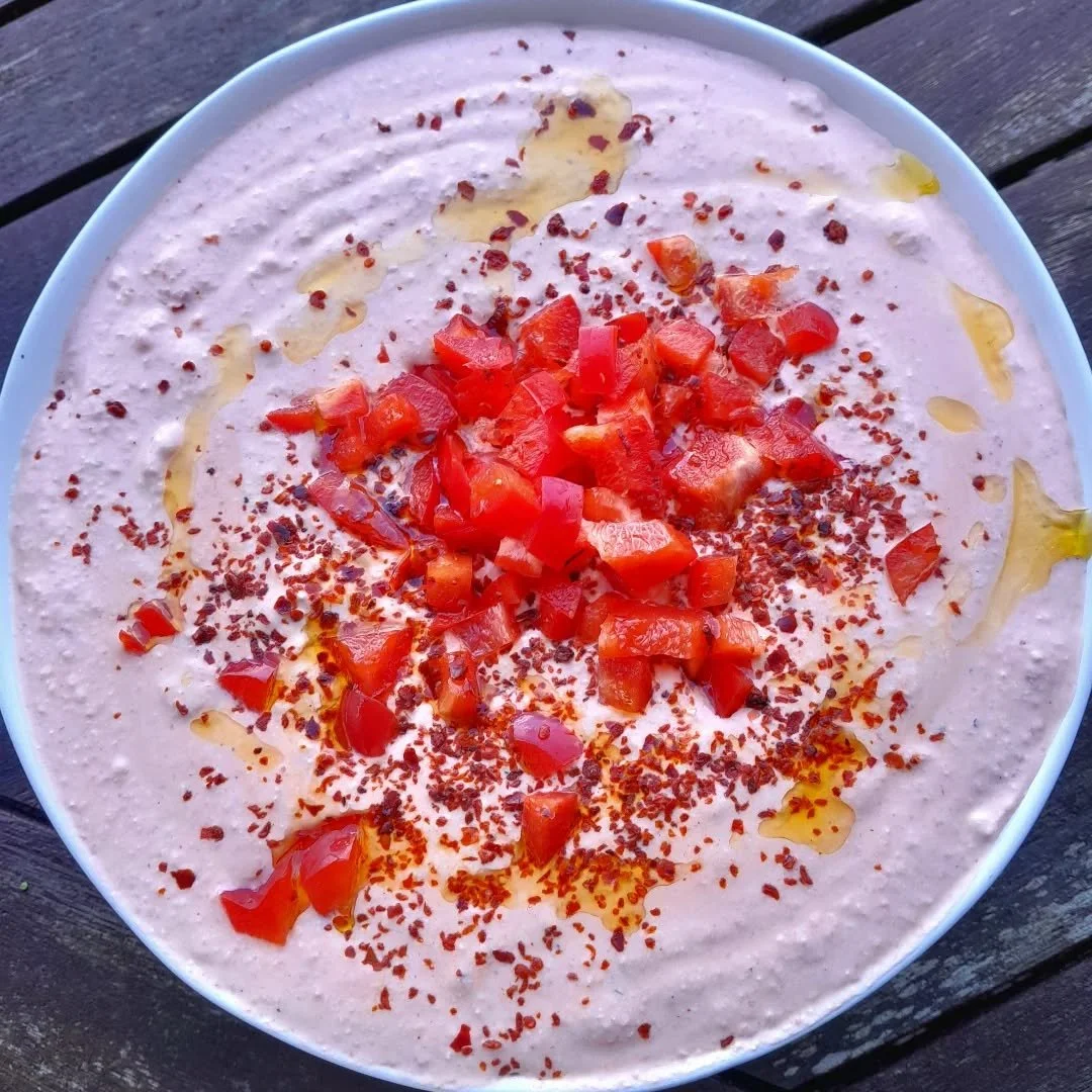 A bowl of creamy, pink-colored dip garnished with chopped red peppers, red pepper flakes, and drizzled with olive oil, placed on a wooden surface.