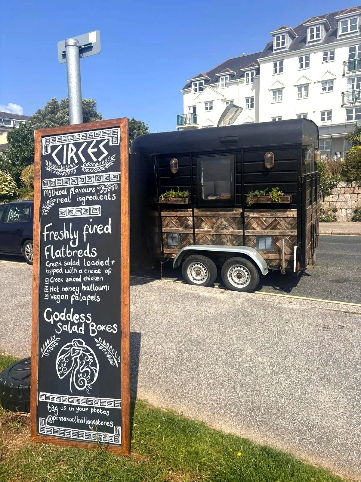 A black food trailer parked on the street with a decorative chalkboard sign in front. The sign advertises a menu of freshly fired flatbreads, Greek salad loaded, Greek spiced chicken, hot honey halloumi, vegan falafels, and goddess salad boxes.