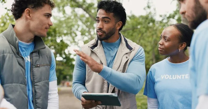 Group of volunteers in conversation outdoors, one man speaking, woman wearing volunteer T-shirt, trees in background.