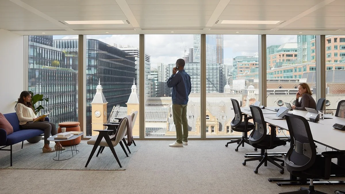 Modern office with large windows overlooking a city skyline, featuring three people: a woman sitting on a couch reading, a man standing and talking on the phone, and a woman at a desk working on a laptop.