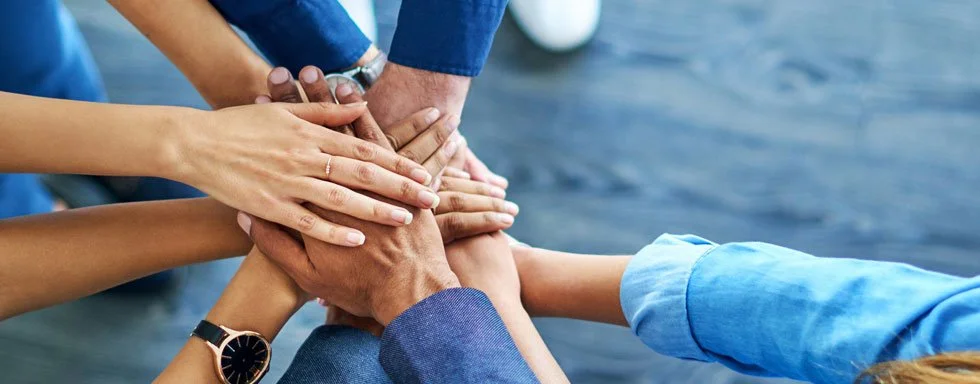 Multiple people placing their hands together in a show of teamwork or unity, with a water body in the background.