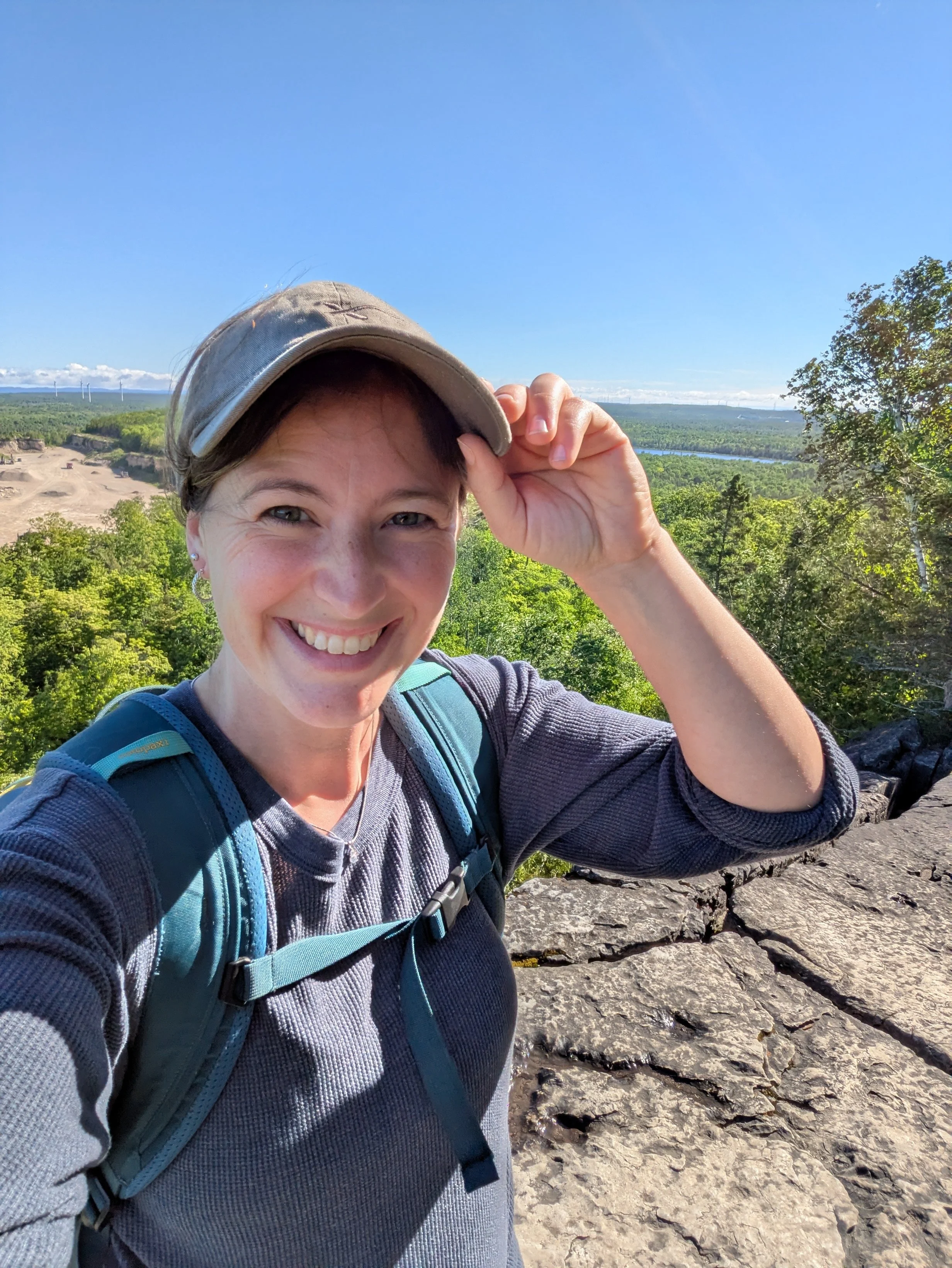 Woman hiking outdoors on a sunny day, wearing a hat and backpack, with a scenic view of green trees, a river, and wind turbines in the background at Belleville Outdoor Preschool