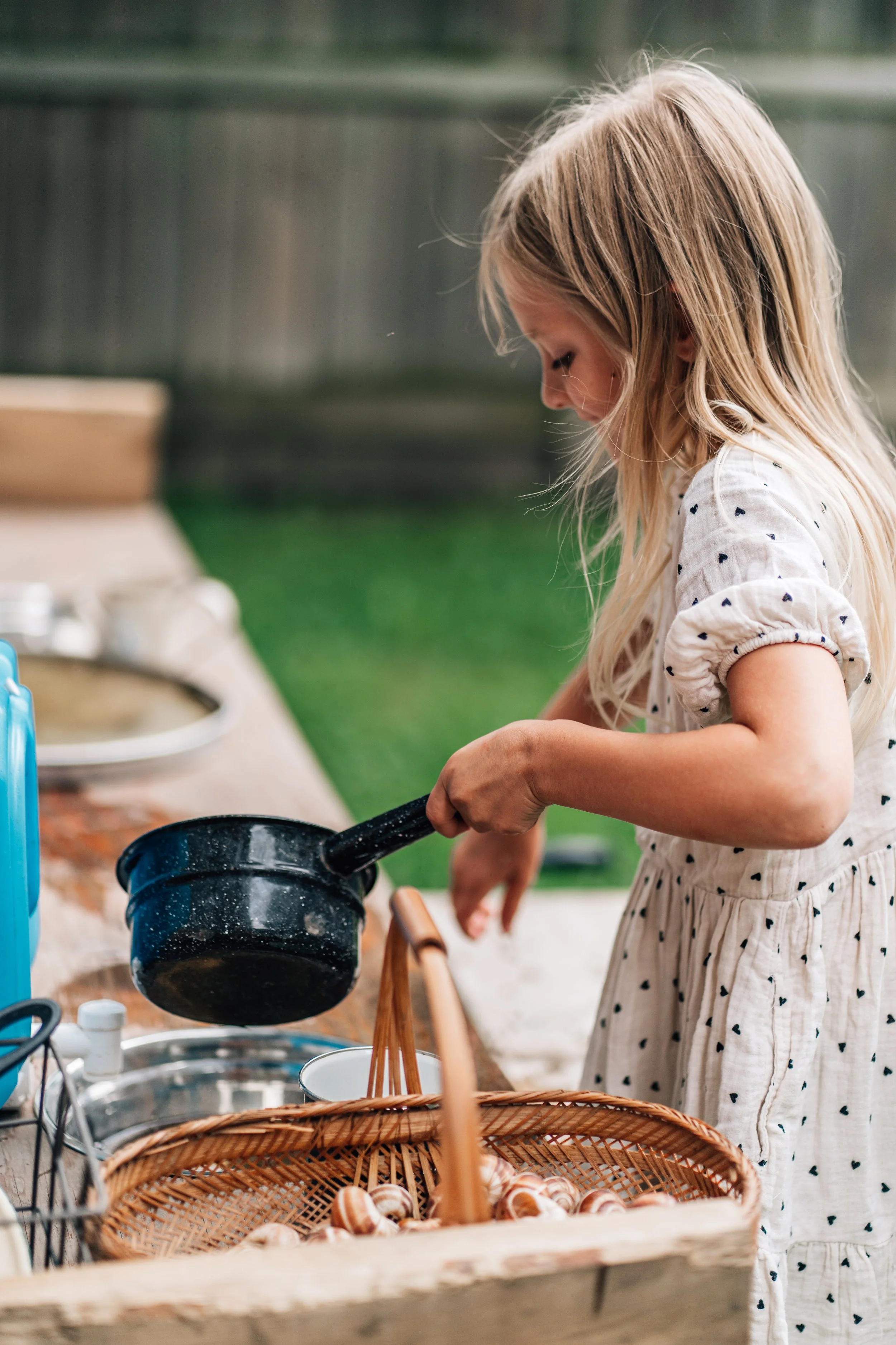 Girl in a white dress pouring water from a black pot into a bowl at the Belleville Outdoor Preschool