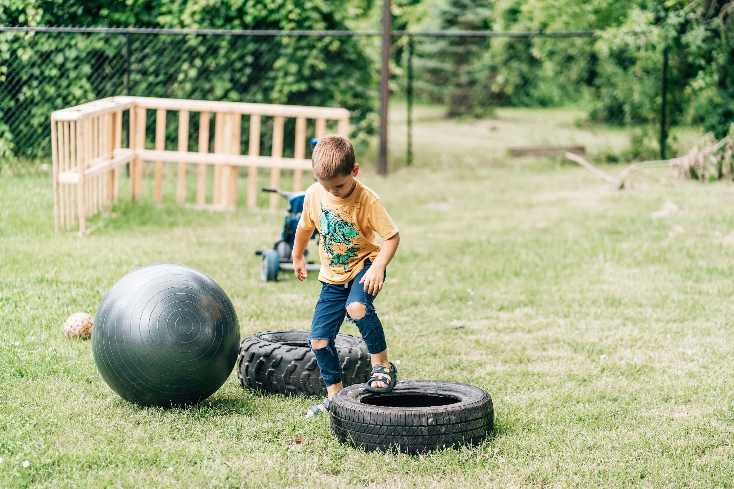 boy in yellow shirt stepping between two tires that are on the grass at the Belleville Outdoor Preschool