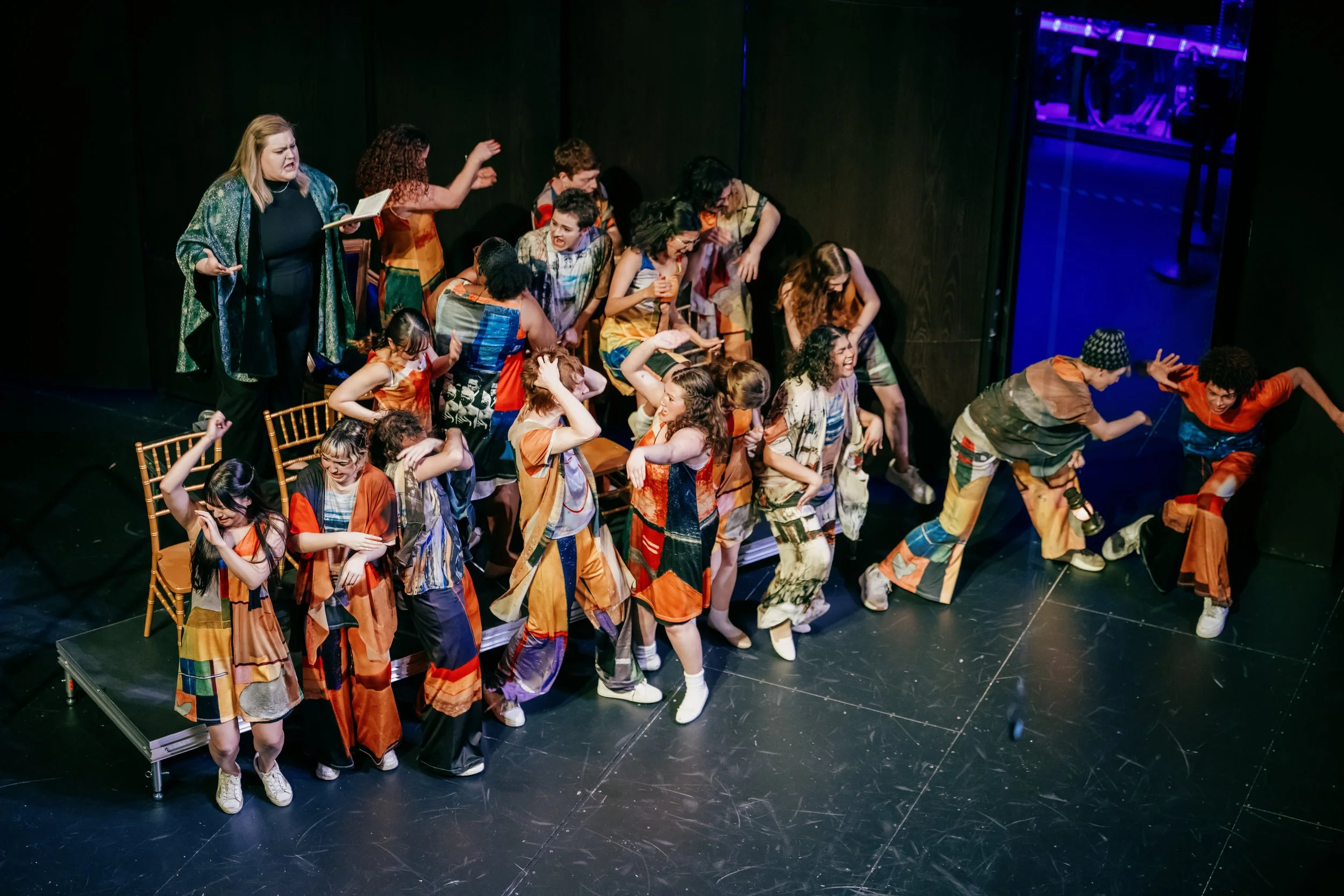 The image shows a group of young women performing on stage, wearing colorful patchwork dresses, with some dancing and others watching. There is an older woman in black with a green patterned shawl standing to the side, holding a script or book. The b