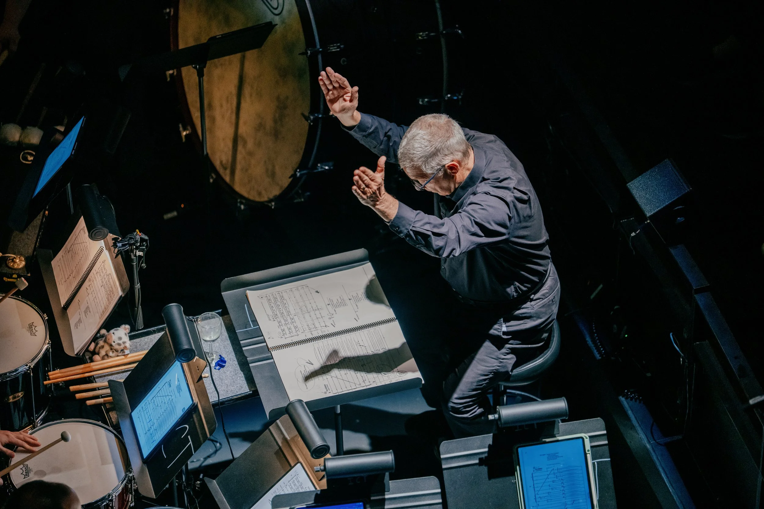 A man with gray hair and glasses, conducting an orchestra or choir, is standing at a music stand with sheet music. Musical instruments, including drums and electronic devices with screens, are visible around him.