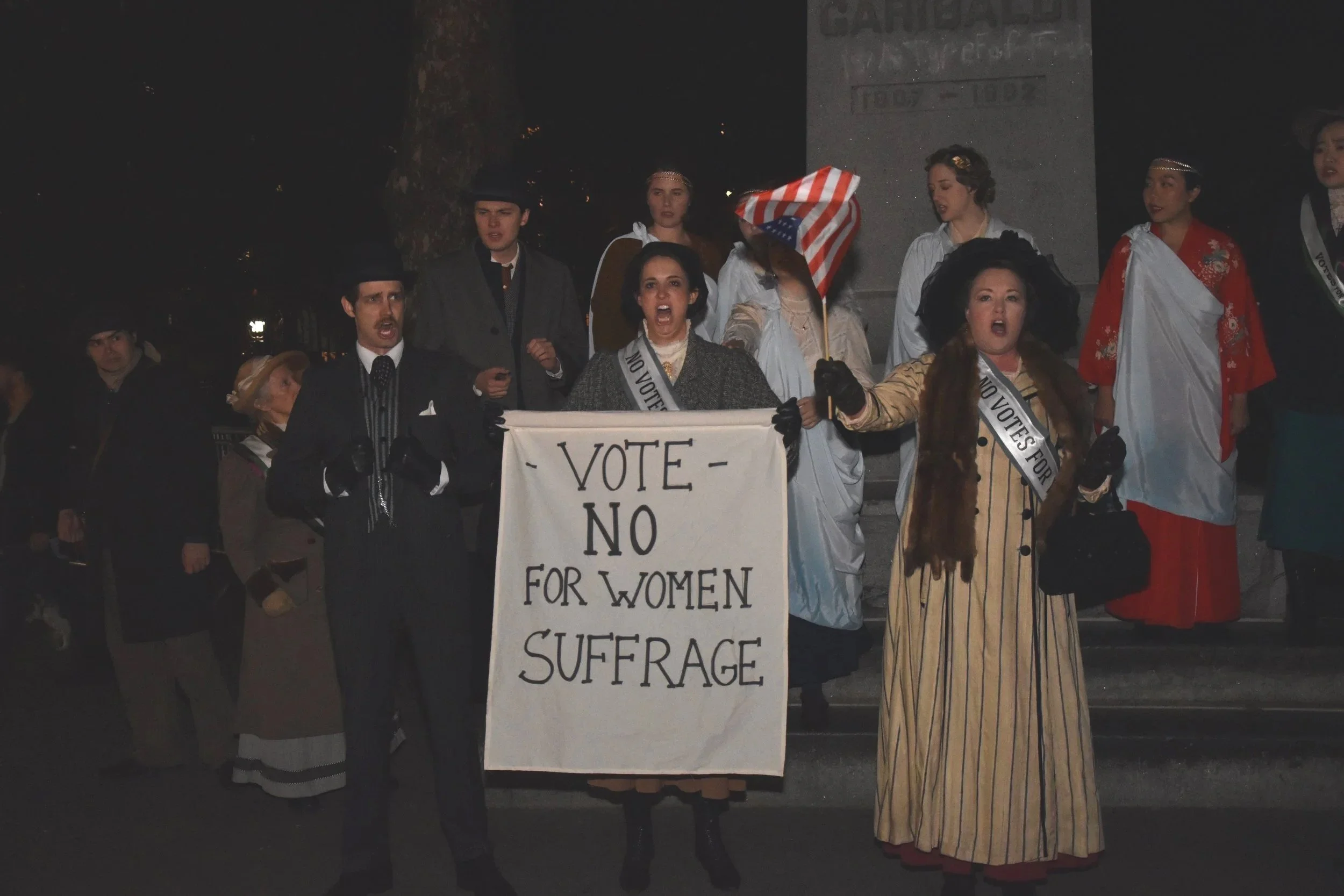 A group of people at night engaged in a protest or demonstration. Some are dressed in historical clothing, holding signs, with one sign reading 'VOTE NO FOR WOMEN'S SUFFRAGE'.