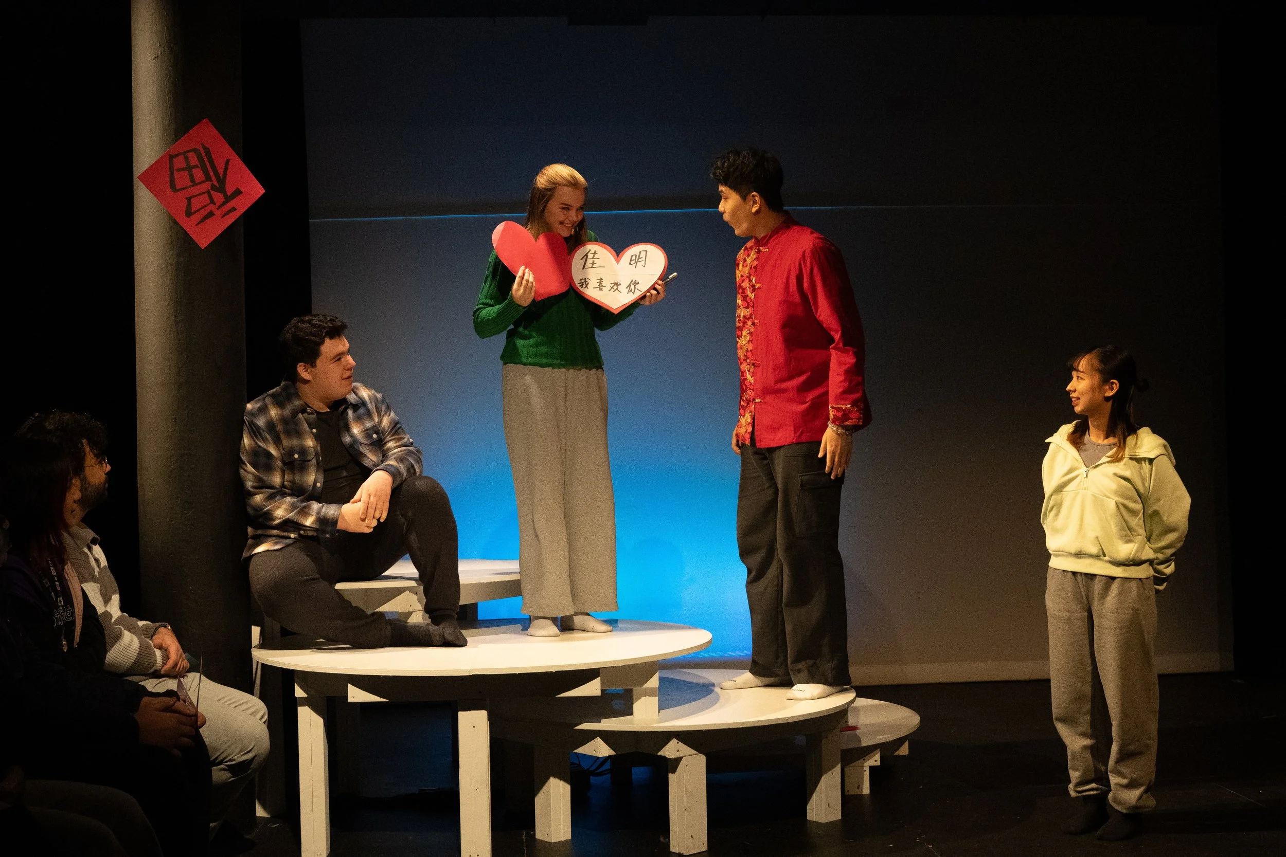 A group of people participating in a theater or stage performance, with some sitting around a circular stage and one standing behind a small elevated platform. The standing woman is holding heart-shaped signs with Chinese characters and smiling at a 