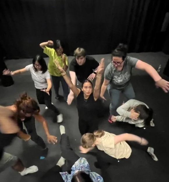 Group of college students, some standing, some sitting or lying on the floor, in a dance or acting studio with black curtains, engaging in a viewpoints theatre workshop.